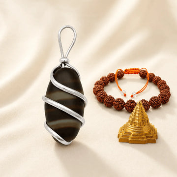 A silver-wrapped black and gray banded stone pendant with a silver bail, displayed next to a brown rudraksha bead bracelet with an orange cord and a gold-toned pyramid pendant, all arranged on a beige satin fabric background.