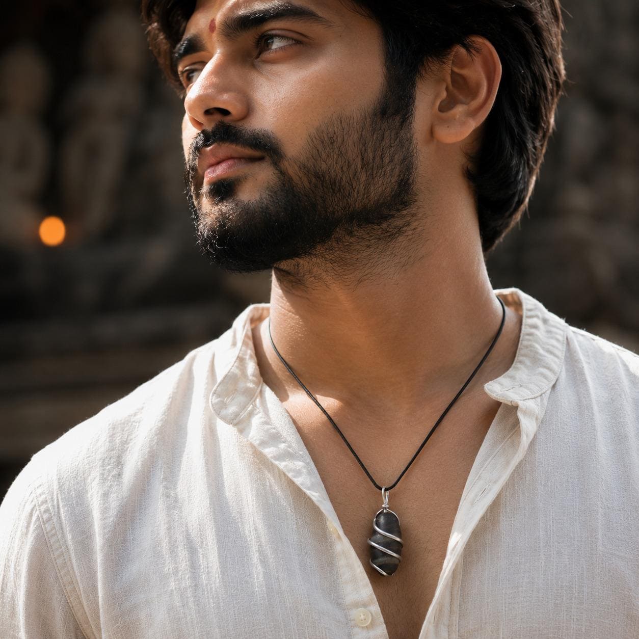 A close-up lifestyle shot of a man wearing a black cord necklace with a dark gray and white striped stone pendant, paired with a white linen shirt.