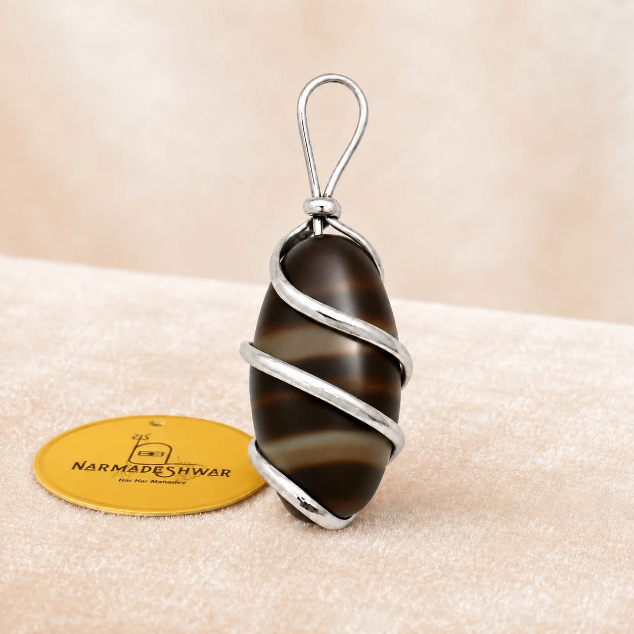 A silver-wrapped pendant featuring a smooth, polished dark brown and black striped stone, displayed next to a yellow circular brand tag on a textured beige surface.